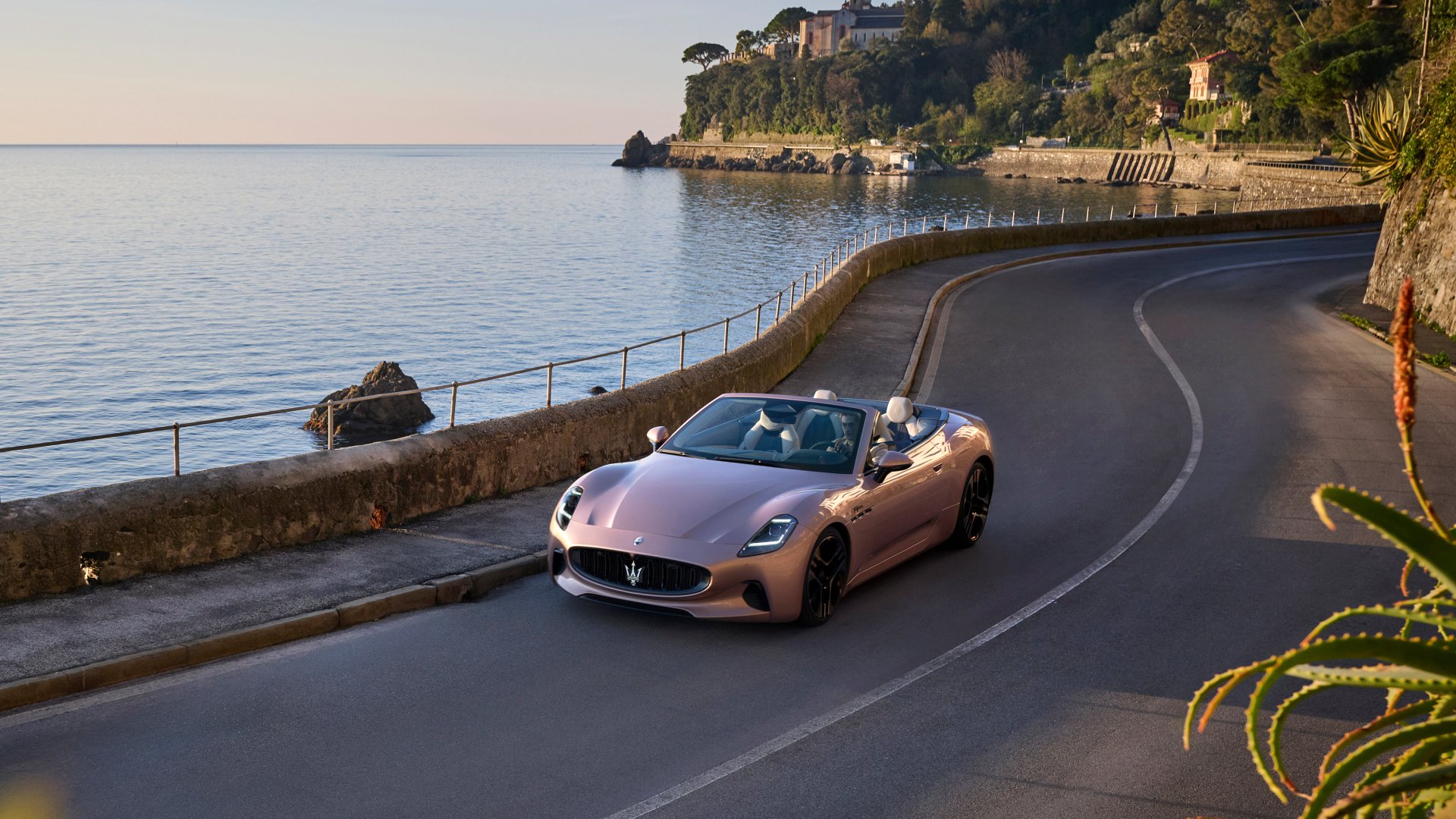 A Maserati GranCabrio drives along a scenic coastal road, with serene waters and lush greenery in the background, creating a stunning HD desktop wallpaper.