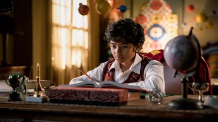 A young boy sits at a desk surrounded by books and a globe, captured in a scene from the TV show *Young Sheldon*, set against a warm, inviting background.