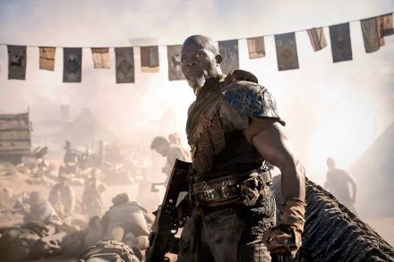 HD wallpaper of a scene from the movie Rebel Moon - Part Two: The Scargiver featuring a focused character in elaborate armor standing strong amidst a dusty, busy campsite with flags fluttering above.
