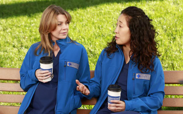 HD wallpaper featuring two characters from the TV show Grey's Anatomy wearing blue scrubs and holding coffee cups while sitting on a bench.
