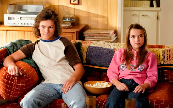 A scene from the TV show Young Sheldon, featuring two young characters seated on a colorful couch, with popcorn in a bowl, set against a cozy background. HD desktop wallpaper.