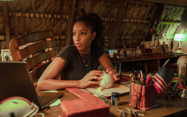 Jessica Williams as a character in the 2024 movie Road House, featured in a HD desktop wallpaper, sitting thoughtfully at a desk with a laptop and various items.