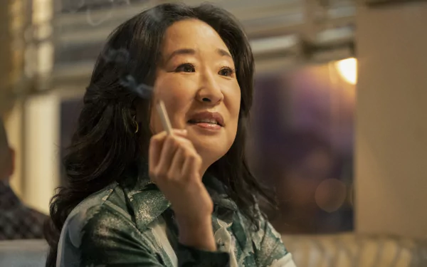 A woman smiles gently while holding a cigarette, dressed in a patterned blouse with soft indoor lighting in the background, from the TV show The Sympathizer. This image is a high-definition desktop wallpaper.