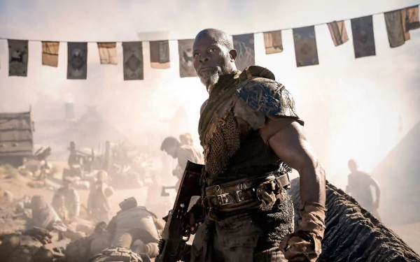 HD wallpaper of a scene from the movie Rebel Moon - Part Two: The Scargiver featuring a focused character in elaborate armor standing strong amidst a dusty, busy campsite with flags fluttering above.
