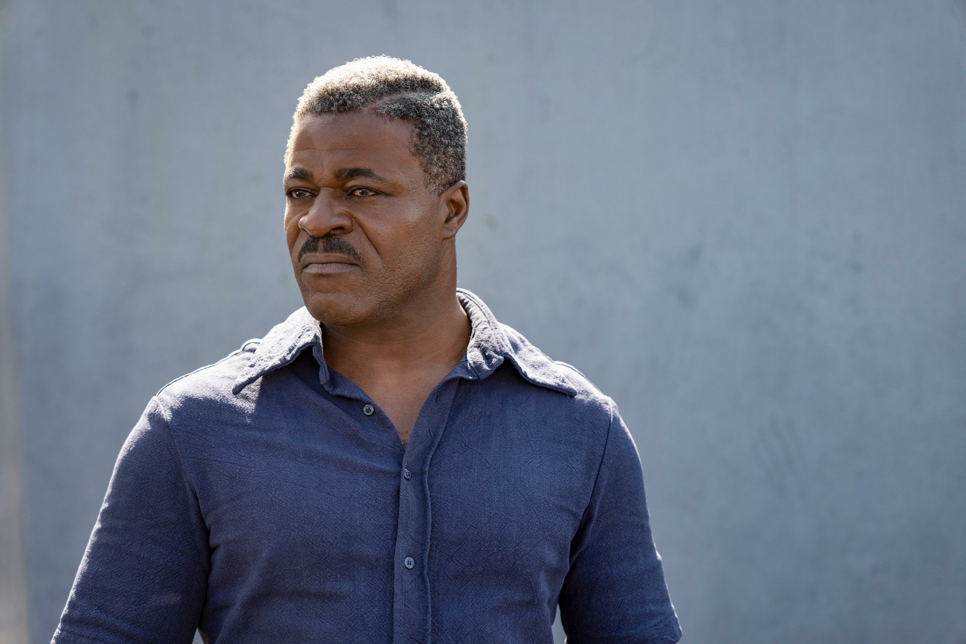 An intense-looking man in a blue shirt stands with a serious expression, possibly a scene from the movie The Ministry of Ungentlemanly Warfare, suitable as an HD desktop wallpaper and background.