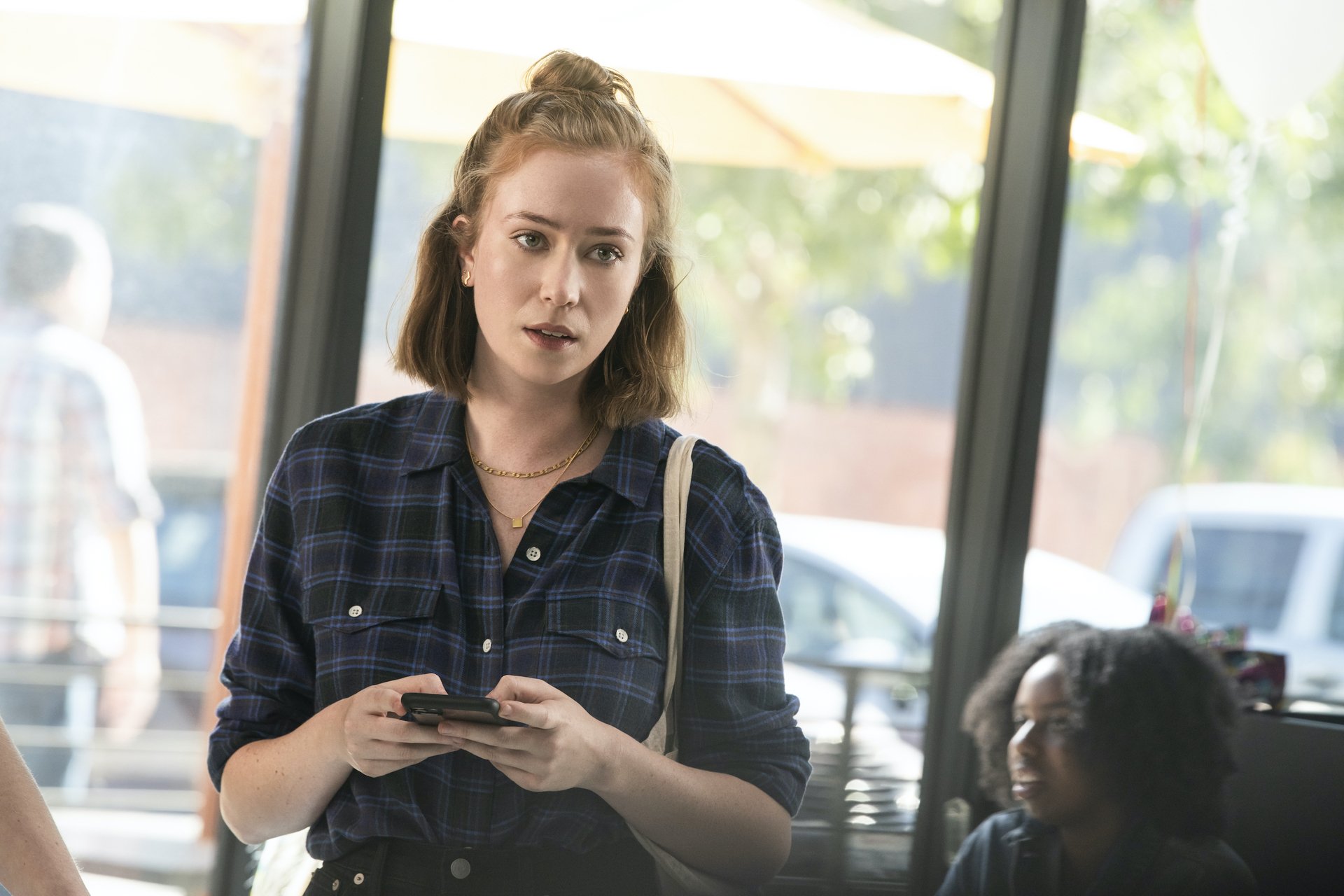 HD desktop wallpaper from the TV show Hacks (2021) featuring a young woman with red hair in a plaid shirt, using her phone in a cafe setting.