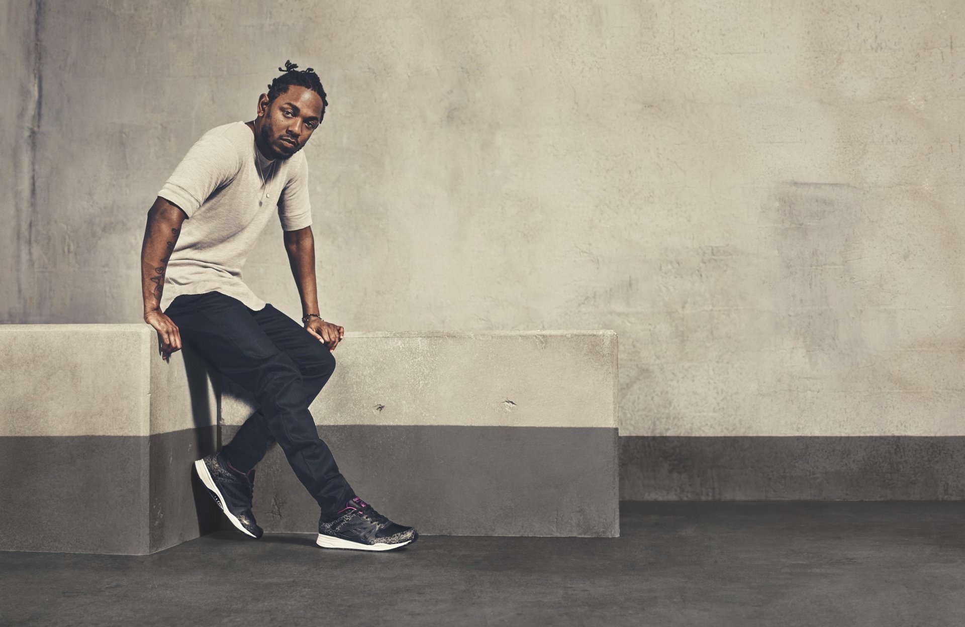 HD desktop wallpaper featuring Kendrick Lamar in a casual pose, dressed in a white t-shirt and black pants, against a textured grey background, embodying a blend of style and music.