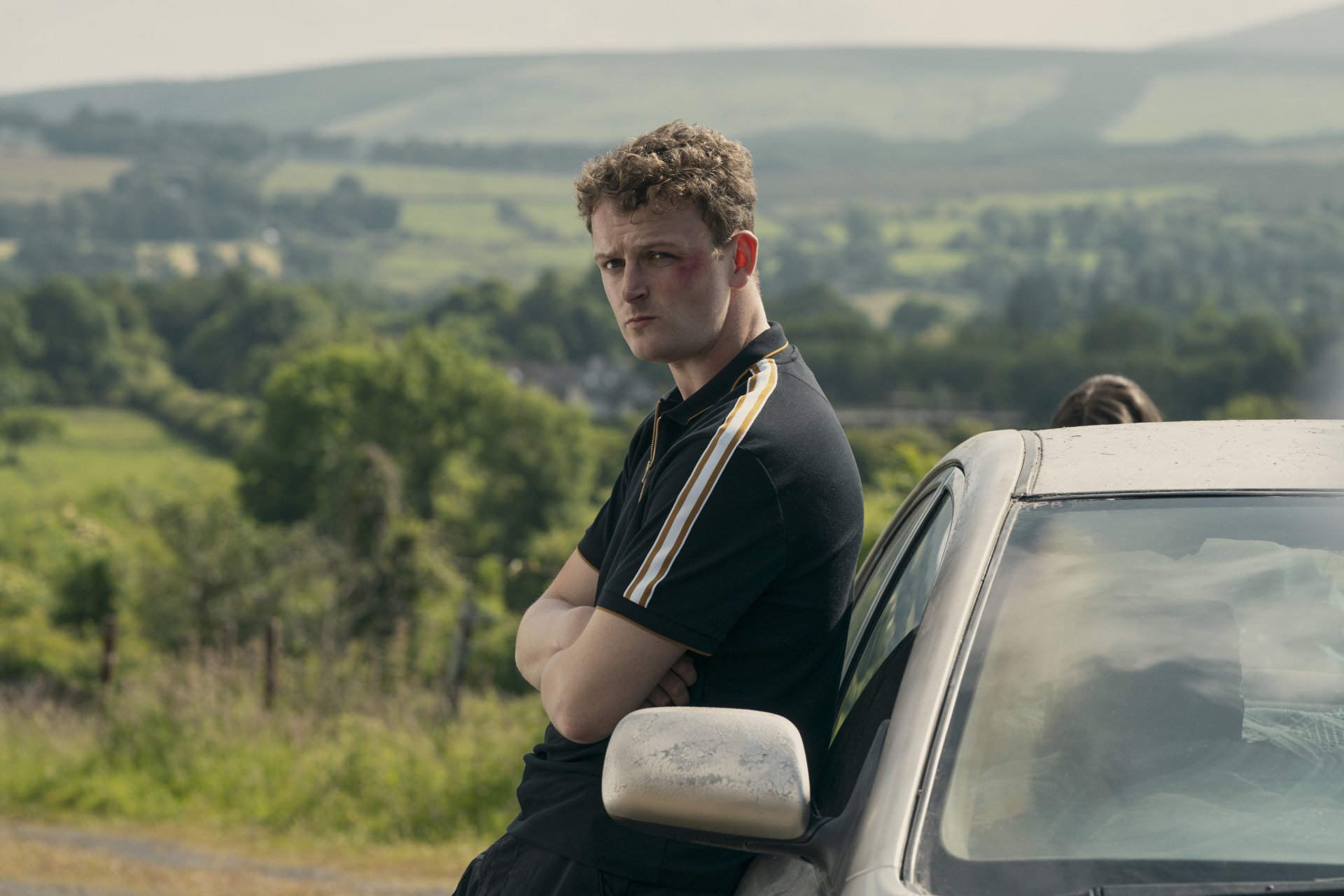 HD wallpaper from the TV show Bodkin featuring a young man leaning on a car with scenic countryside in the background.