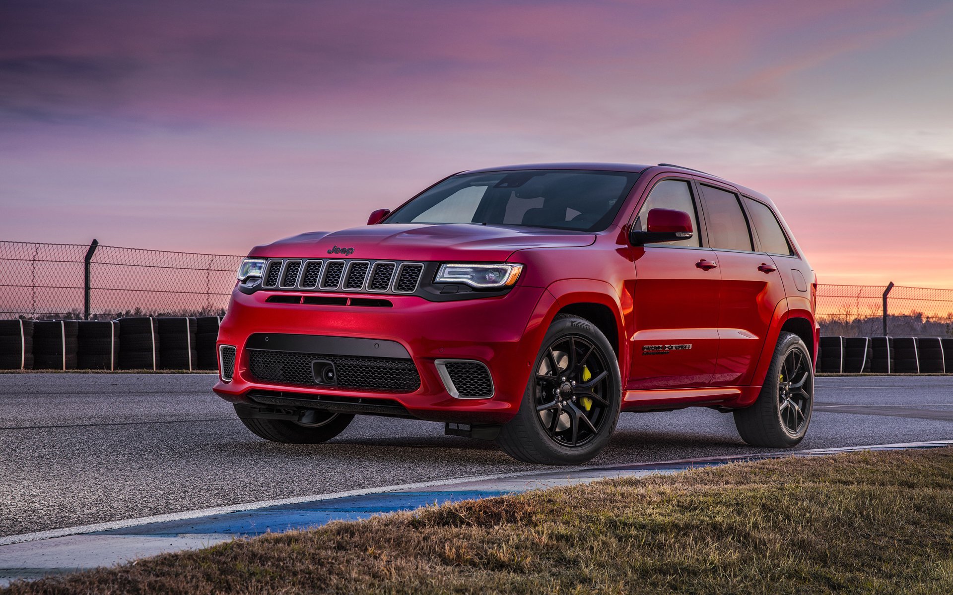 HD desktop wallpaper featuring a striking red Jeep Grand Cherokee parked on a tarmac, with a fenced racetrack and a colorful sunset sky in the background.