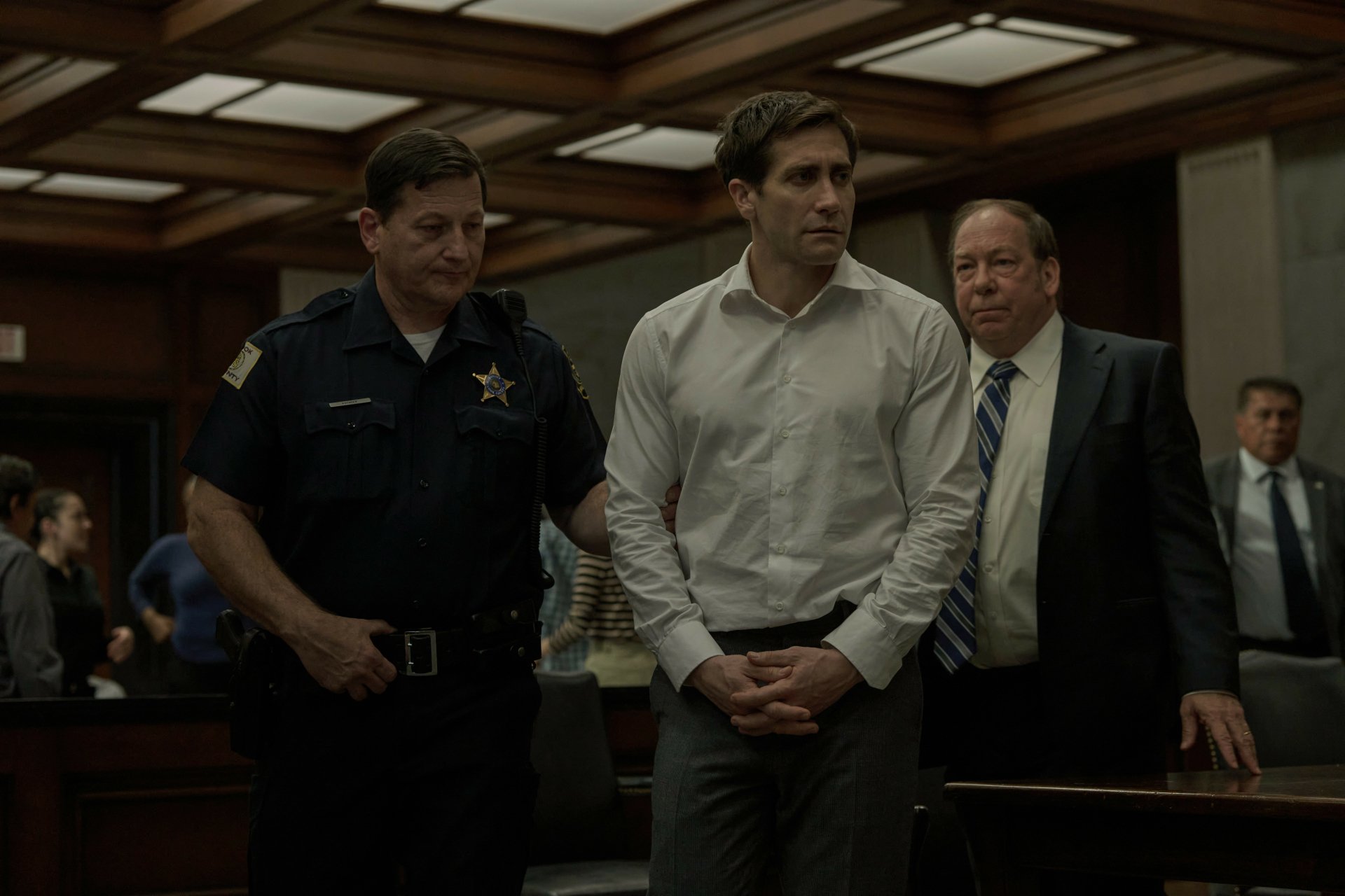 HD wallpaper from the TV show Presumed Innocent (2024), featuring Jake Gyllenhaal in a courtroom setting, flanked by a police officer and a man in a suit.