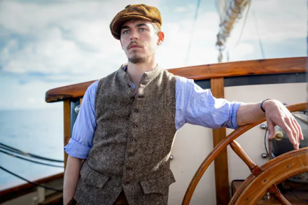 A man in period clothing stands by a ship's wheel with the ocean in the background, from the movie The Ministry of Ungentlemanly Warfare, suitable as an HD desktop wallpaper.