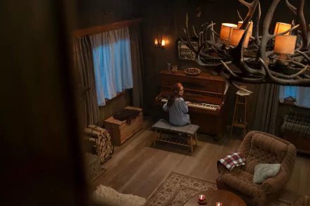 A high-definition wallpaper from The Strangers: Chapter 1, featuring Madelaine Petsch playing piano in a cozy, dimly lit cabin with rustic decor.