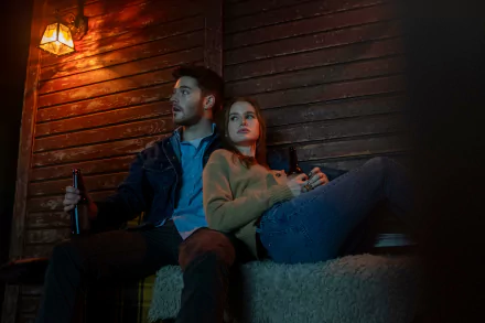 HD desktop wallpaper of The Strangers: Chapter 1 featuring Madelaine Petsch sitting with a man in a dimly lit, rustic setting, both holding bottles, creating a suspenseful atmosphere.