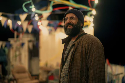 HD wallpaper featuring a scene from the TV show Sweet Tooth, showcasing a bearded man wearing a hat and glasses at a colorful, festive outdoor setting with lights and bunting in the background.