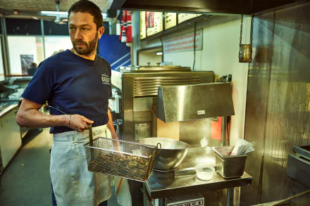 HD desktop wallpaper of Ebon Moss-Bachrach as a character from the TV show The Bear, standing in a kitchen, holding a fryer basket, with kitchen equipment in the background.