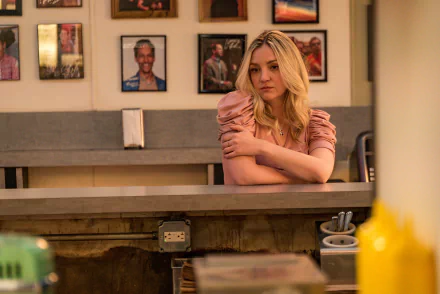 HD desktop wallpaper of Abby Elliott in a scene from the TV show The Bear, featuring her sitting behind a diner counter with framed photos on the wall.