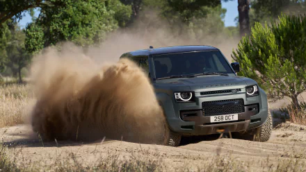 A Land Rover Defender Octa driving through sandy terrain, kicking up a cloud of sand. The HD desktop wallpaper showcases the vehicle's rugged design amidst a background of trees and vegetation.