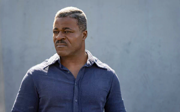 An intense-looking man in a blue shirt stands with a serious expression, possibly a scene from the movie The Ministry of Ungentlemanly Warfare, suitable as an HD desktop wallpaper and background.