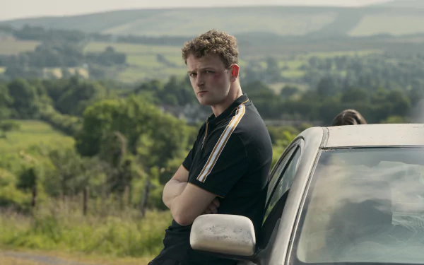 HD wallpaper from the TV show Bodkin featuring a young man leaning on a car with scenic countryside in the background.