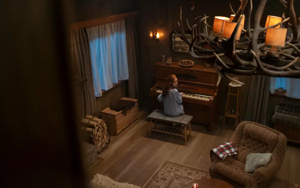 A high-definition wallpaper from The Strangers: Chapter 1, featuring Madelaine Petsch playing piano in a cozy, dimly lit cabin with rustic decor.