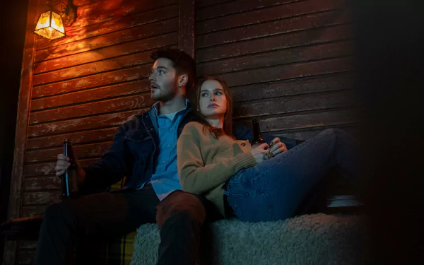 HD desktop wallpaper of The Strangers: Chapter 1 featuring Madelaine Petsch sitting with a man in a dimly lit, rustic setting, both holding bottles, creating a suspenseful atmosphere.