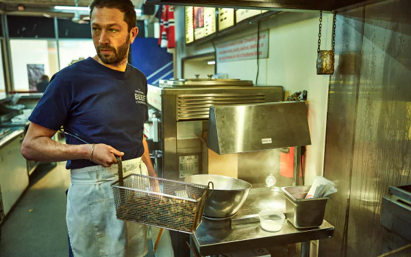 HD desktop wallpaper of Ebon Moss-Bachrach as a character from the TV show The Bear, standing in a kitchen, holding a fryer basket, with kitchen equipment in the background.