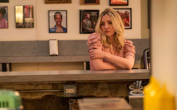 HD desktop wallpaper of Abby Elliott in a scene from the TV show The Bear, featuring her sitting behind a diner counter with framed photos on the wall.