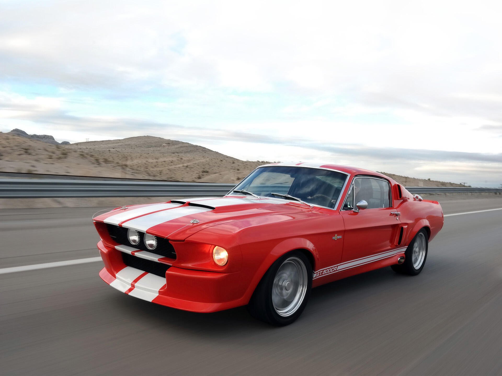 HD desktop wallpaper of a classic red Shelby GT500CR car with white racing stripes driving on a road near a desert landscape.