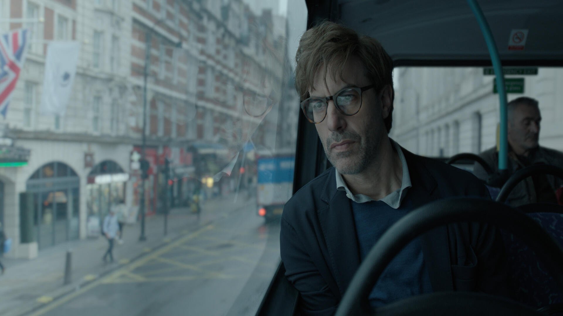 A 4K Ultra HD wallpaper featuring a scene from the TV show Disclaimer (2024), showcasing Sacha Baron Cohen looking pensively from a bus amid a rainy city backdrop.