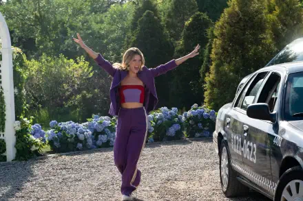 Meghann Fahy joyfully runs towards a taxi in this vibrant scene from the TV show The Perfect Couple, surrounded by lush greenery and colorful flowers.