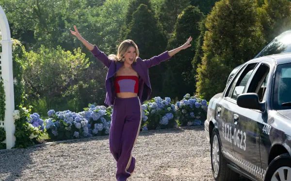 Meghann Fahy joyfully runs towards a taxi in this vibrant scene from the TV show The Perfect Couple, surrounded by lush greenery and colorful flowers.