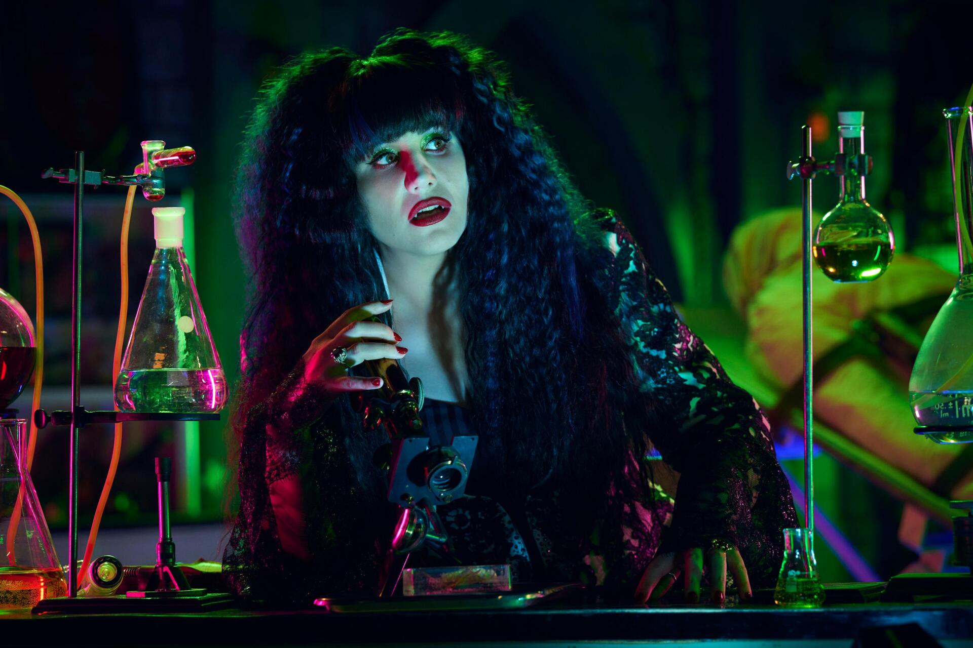 Natasia Demetriou in a scene from What We Do in the Shadows, featuring a vibrant laboratory setting with colorful lighting and intricate glassware. HD wallpaper background for fans.