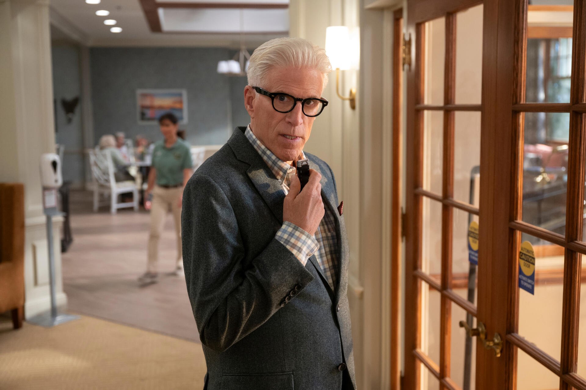 A scene from the TV show A Man on the Inside, featuring Ted Danson in a stylish suit, contemplating in a well-lit room with a glimpse of another character in the background.