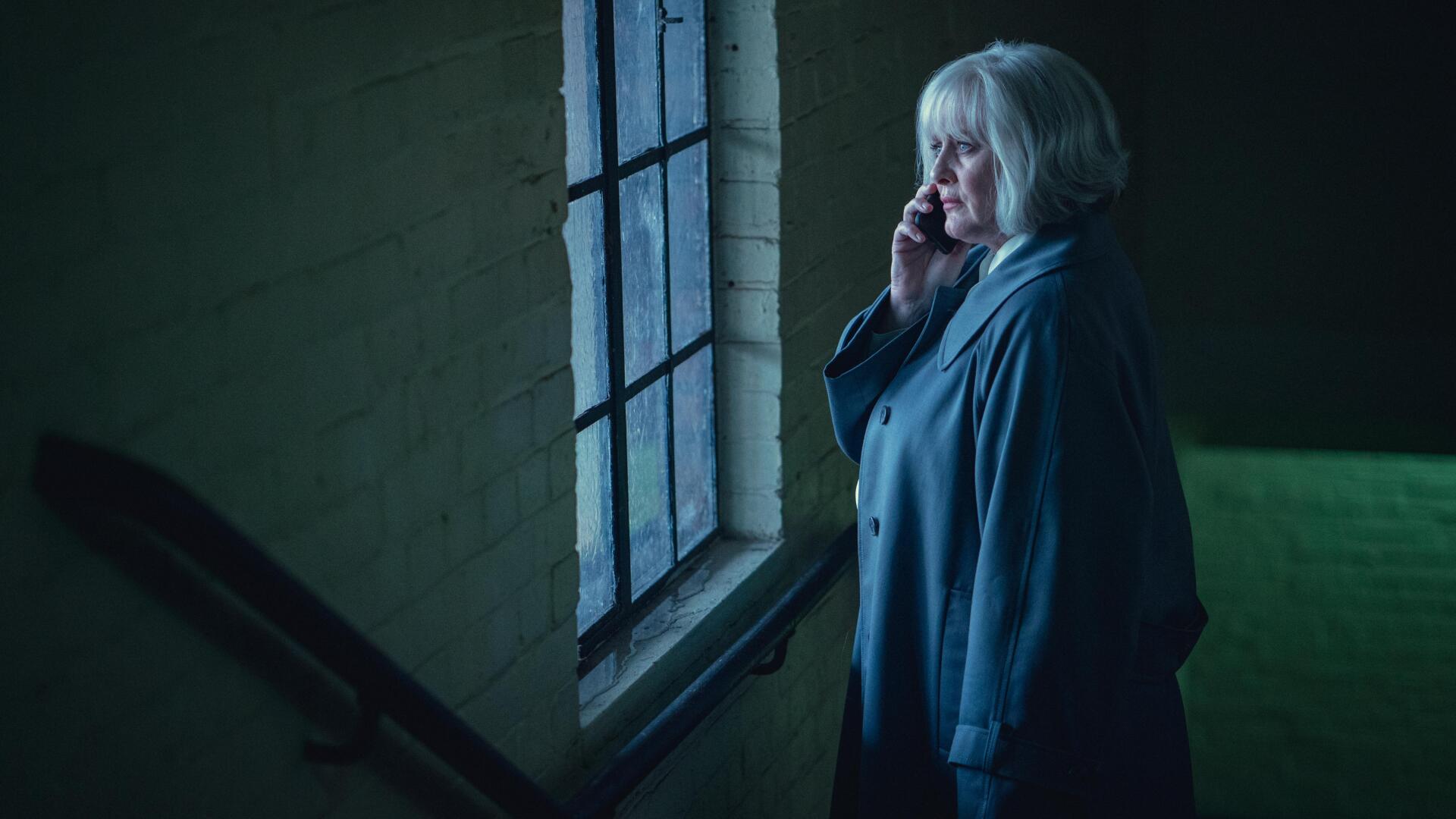 A scene from the TV show Black Doves featuring a woman with short gray hair speaking on the phone by a window, captured in atmospheric lighting. 4K Ultra HD wallpaper and background.