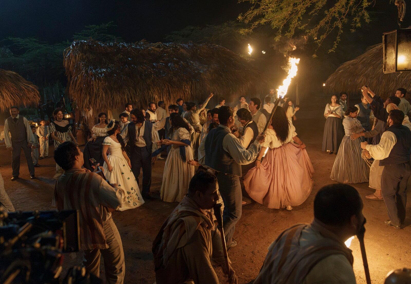 A vibrant scene from the TV show One Hundred Years of Solitude, depicting a lively celebration with characters dancing around fires in a rustic setting. HD desktop wallpaper.