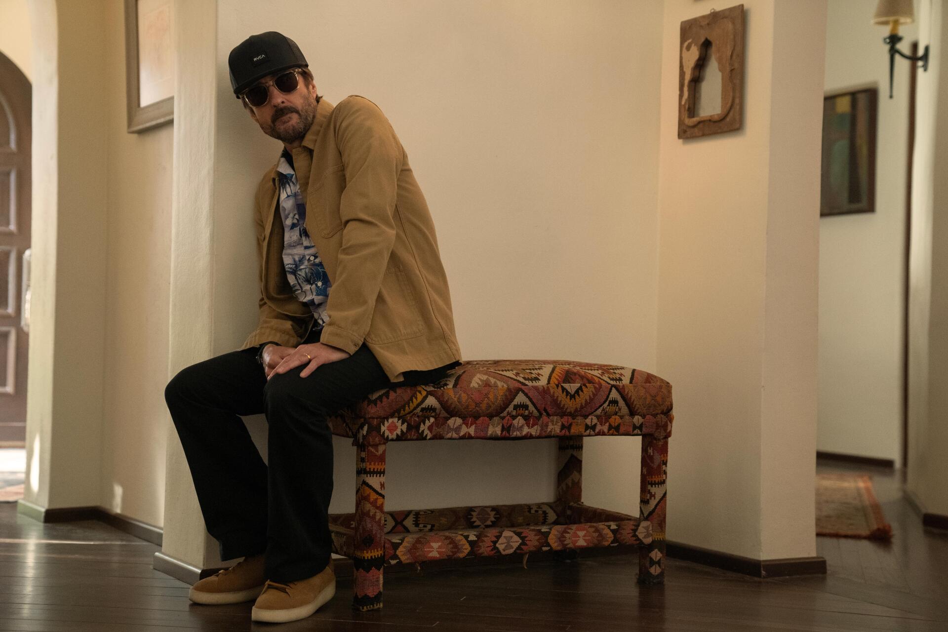 A scene from the 2024 TV show No Good Deed features Luke Wilson sitting on a decorative bench, dressed casually with sunglasses, set in a stylish indoor environment.
