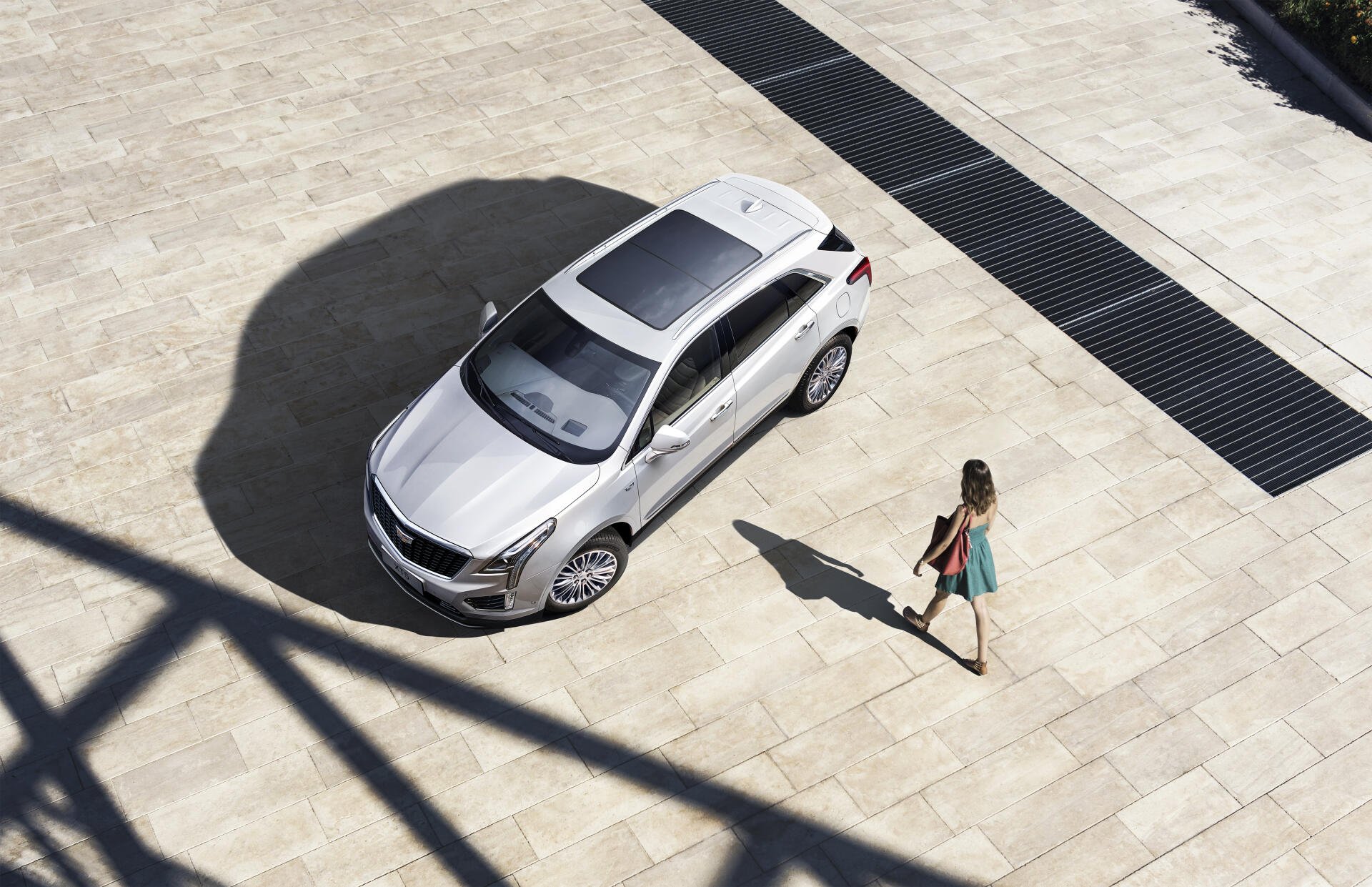 A sleek silver Cadillac XT5 parked on a sunlit stone surface, with a woman in a colorful dress walking nearby, creating a modern and stylish scene. 4K Ultra HD quality.