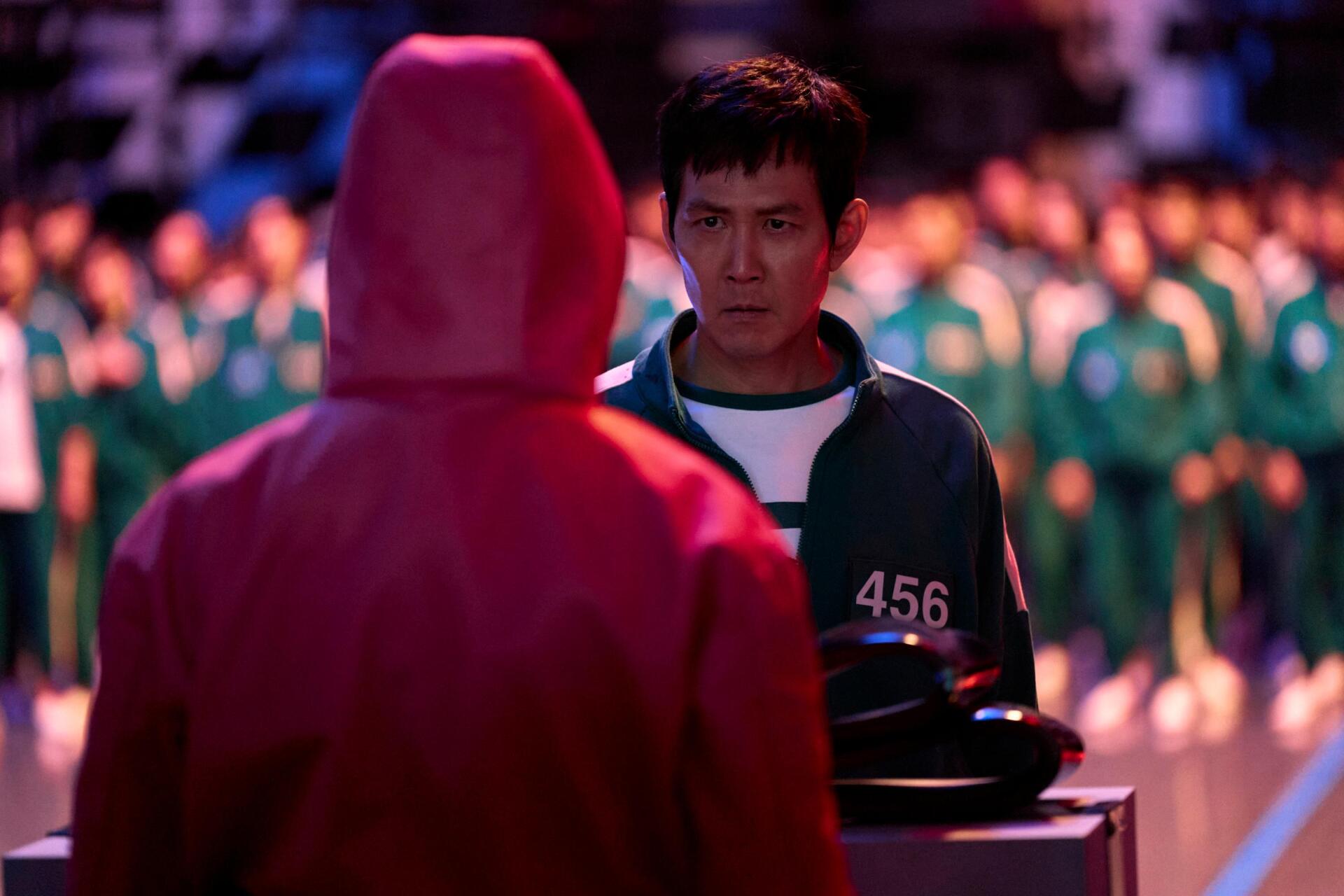 A scene from Squid Game featuring Lee Jung-jae, facing a masked figure in a dramatic moment, surrounded by contestants in green uniforms. This HD image serves as a striking desktop wallpaper.