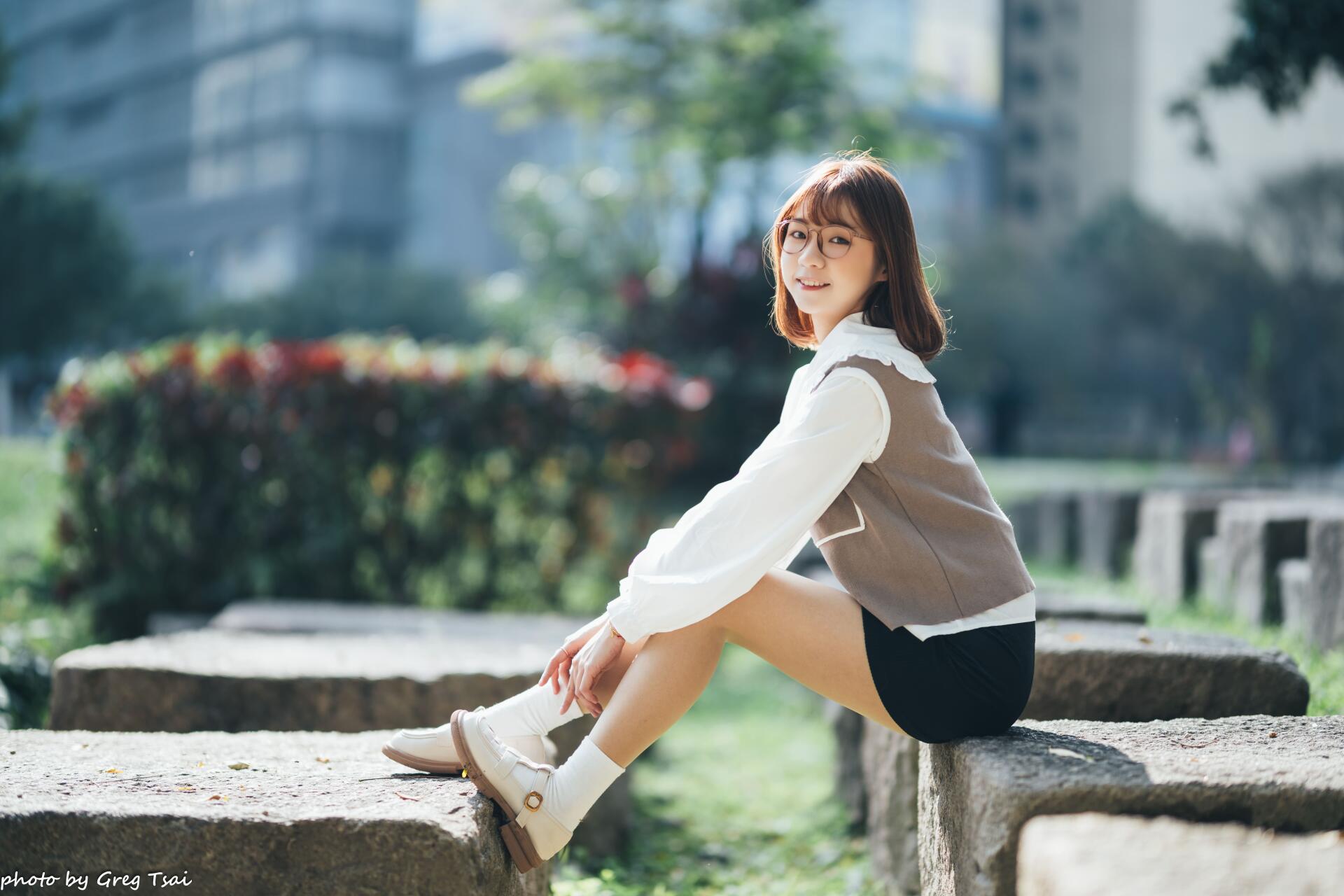 Asian woman seated on a stone ledge in a sunlit park, smiling at the camera with a blurred cityscape behind her — 8K Ultra HD PC desktop wallpaper and background.