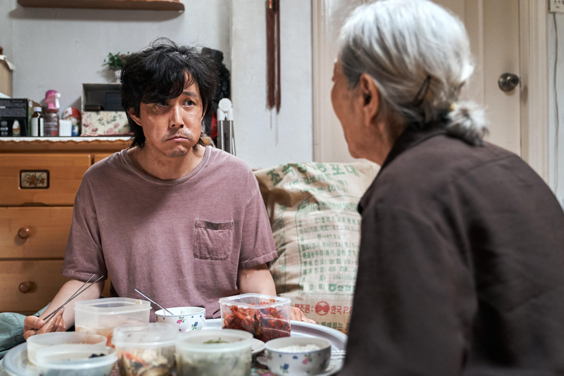 A scene from the TV show Squid Game featuring Lee Jung-jae, sitting at a table with dishes, engaged in a conversation with an elderly woman. Vibrant 4K Ultra HD wallpaper.