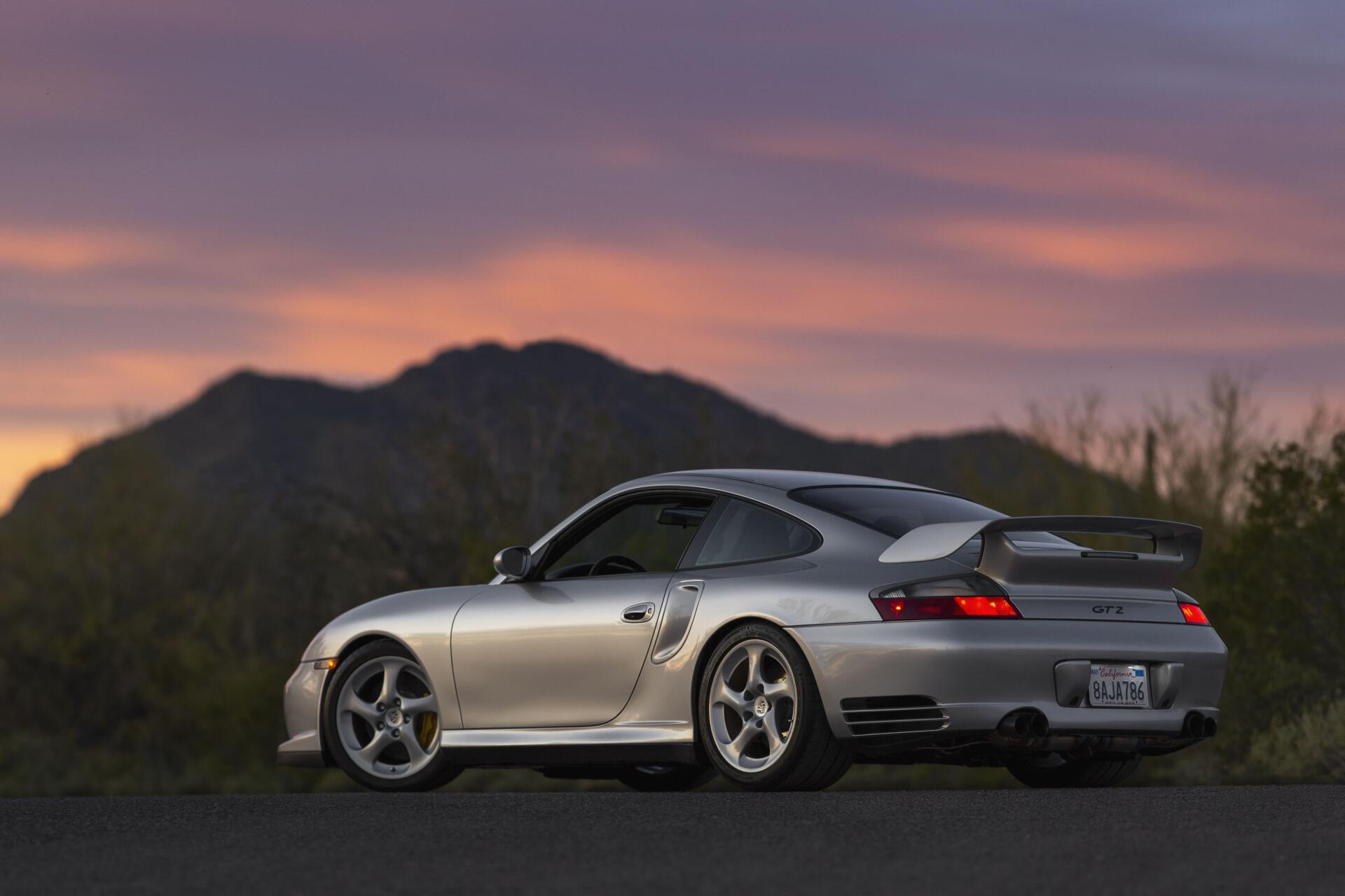 4K Ultra HD PC desktop wallpaper of a silver Porsche parked on a mountain road at sunset, silhouetted hills beneath a purple-orange sky.