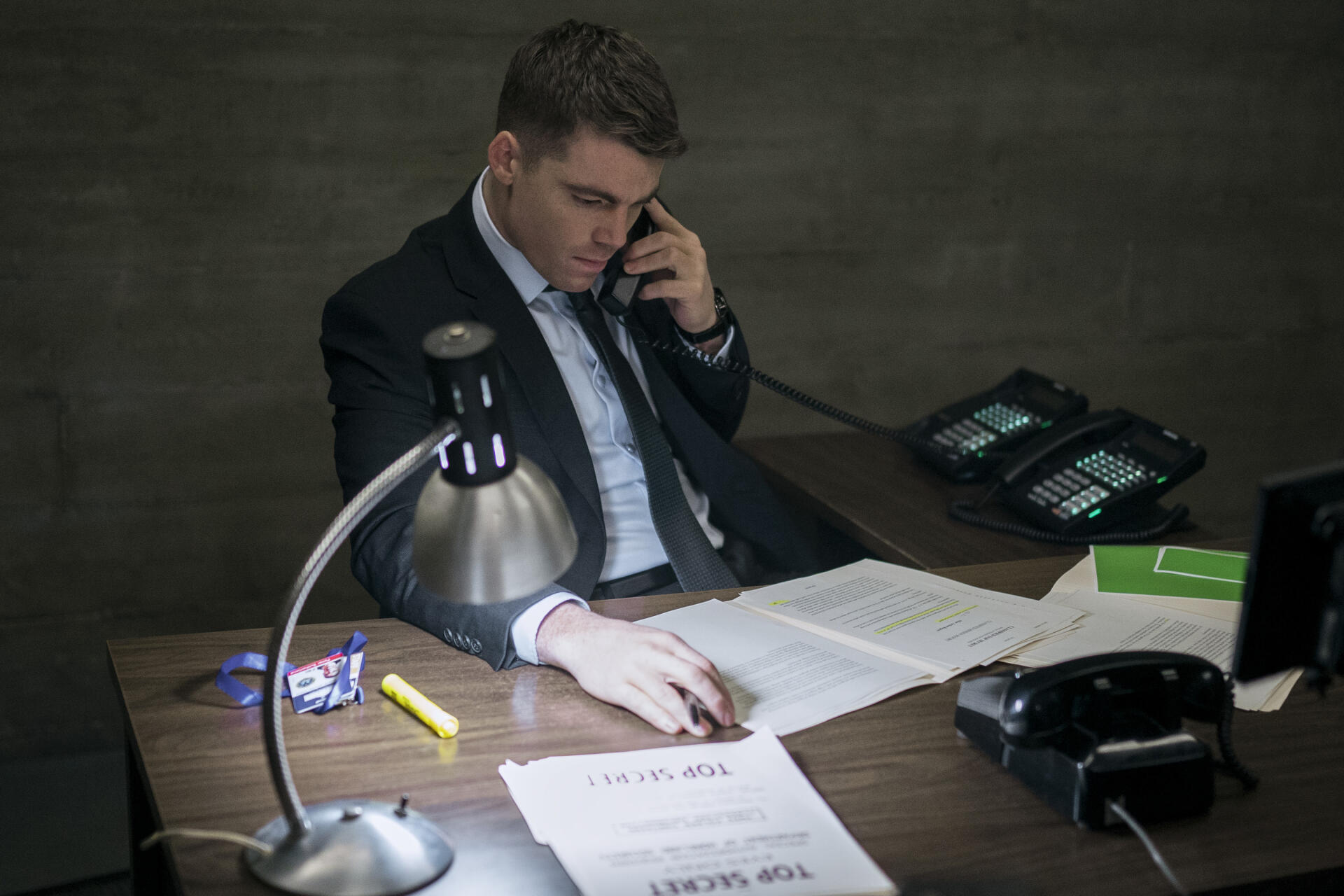 A scene from the TV show The Night Agent featuring Gabriel Basso seated at a desk, talking on the phone with documents spread out, illuminated by a desk lamp.