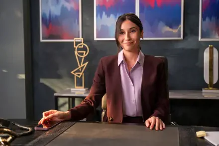 A confident woman sits at a desk in an office, smiling as she engages with her surroundings. The backdrop features abstract art, representing the TV show A Man on the Inside.