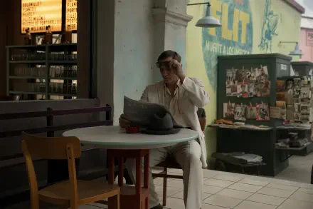 A stylish Daniel Craig reading a newspaper at a café, surrounded by an artistic urban backdrop, featured in a movie with queer themes. This 4K Ultra HD image serves as a captivating desktop wallpaper.