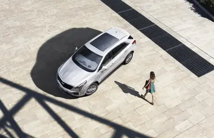 A sleek silver Cadillac XT5 parked on a sunlit stone surface, with a woman in a colorful dress walking nearby, creating a modern and stylish scene. 4K Ultra HD quality.