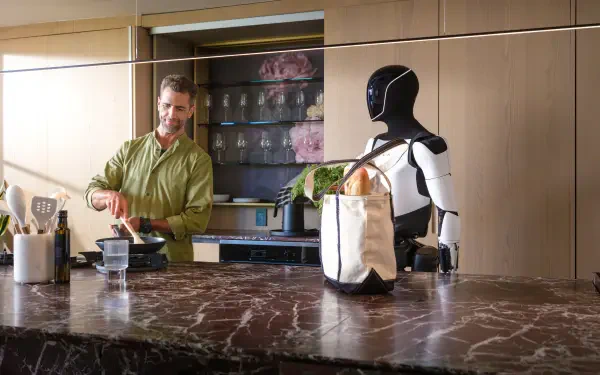 A man cooks in a modern kitchen while the Tesla Optimus robot, a futuristic assistant, stands beside him. This 4K Ultra HD wallpaper showcases advanced technology in everyday life.