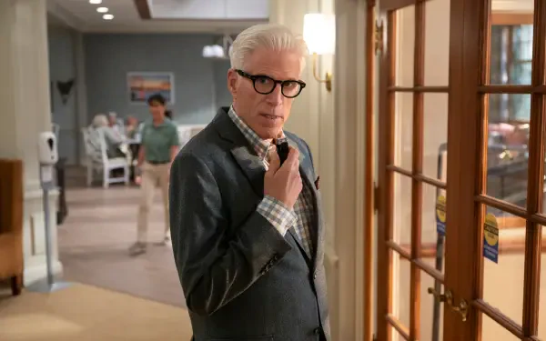A scene from the TV show A Man on the Inside, featuring Ted Danson in a stylish suit, contemplating in a well-lit room with a glimpse of another character in the background.