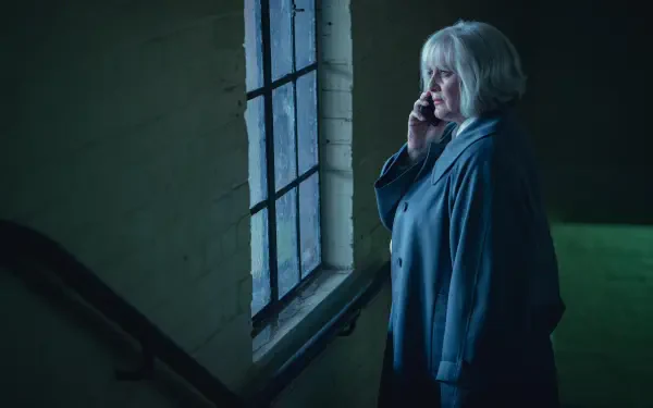 A scene from the TV show Black Doves featuring a woman with short gray hair speaking on the phone by a window, captured in atmospheric lighting. 4K Ultra HD wallpaper and background.