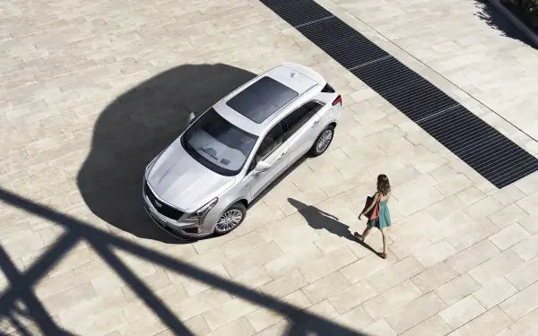 A sleek silver Cadillac XT5 parked on a sunlit stone surface, with a woman in a colorful dress walking nearby, creating a modern and stylish scene. 4K Ultra HD quality.