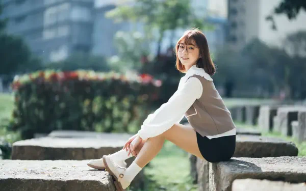 Asian woman seated on a stone ledge in a sunlit park, smiling at the camera with a blurred cityscape behind her — 8K Ultra HD PC desktop wallpaper and background.